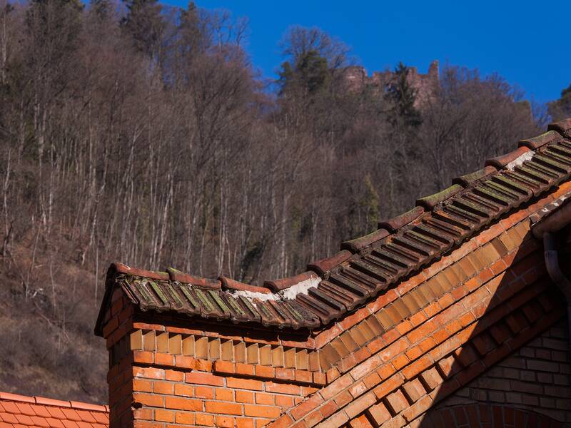 Ein schräges Ziegeldach der Szene 64 wird vor einem bewaldeten Hügel und klarem blauen Himmel gezeigt.